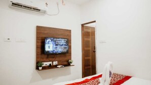 Une chambre d’hôtel avec un lit, un téléviseur fixé au mur, une porte en bois et une petite étagère sur laquelle se trouve une plante.