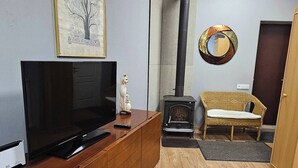Una sala de estar con televisión sobre un mueble de madera, chimenea, una silla de mimbre y un cuadro en la pared.