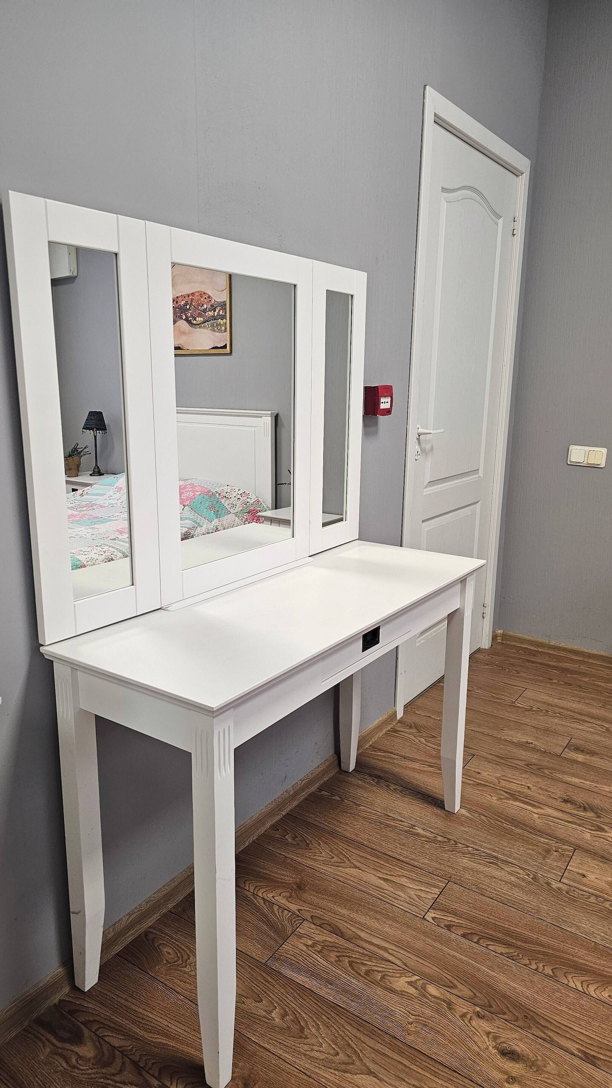 Un comodino bianco con uno specchio, una lampada da comodino piccola e un quadro appeso alla parete.