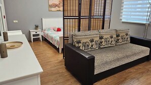 Una habitación con suelo de madera, un escritorio blanco, una cama con colcha estampada, un sofá negro con cojines decorados y un separador de madera, además de una ventana con persianas.