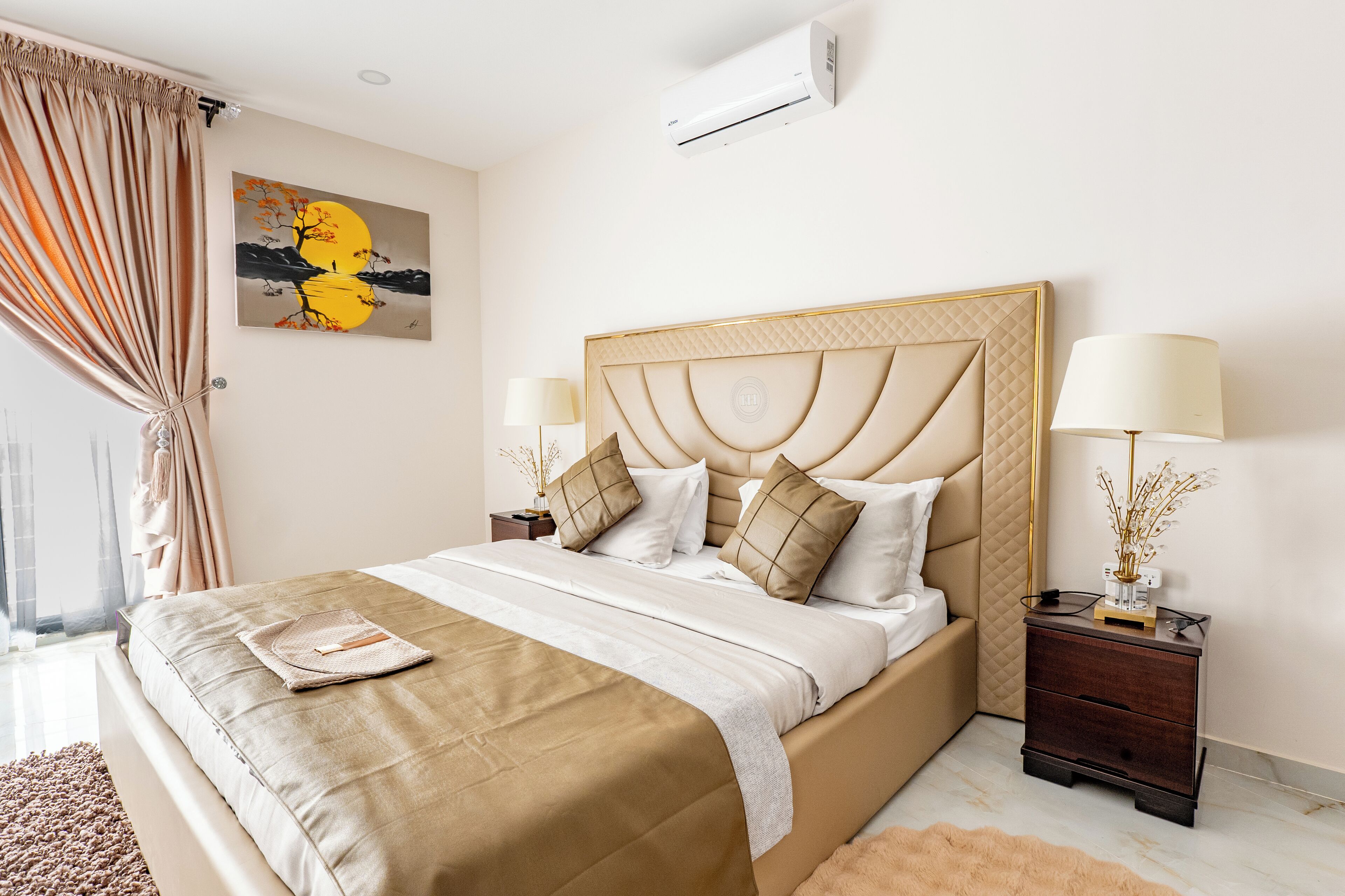 A bedroom with a large bed, two bedside tables, a wall-mounted air conditioner, and a painting.