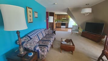 A living room with a sofa, a coffee table, a television, and a lamp. There is a kitchen in the background.