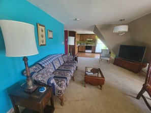 Ein Wohnzimmer mit einer Couch, einem Couchtisch, einem Fernseher und einer Lampe. Im Hintergrund ist eine Küche zu sehen.
