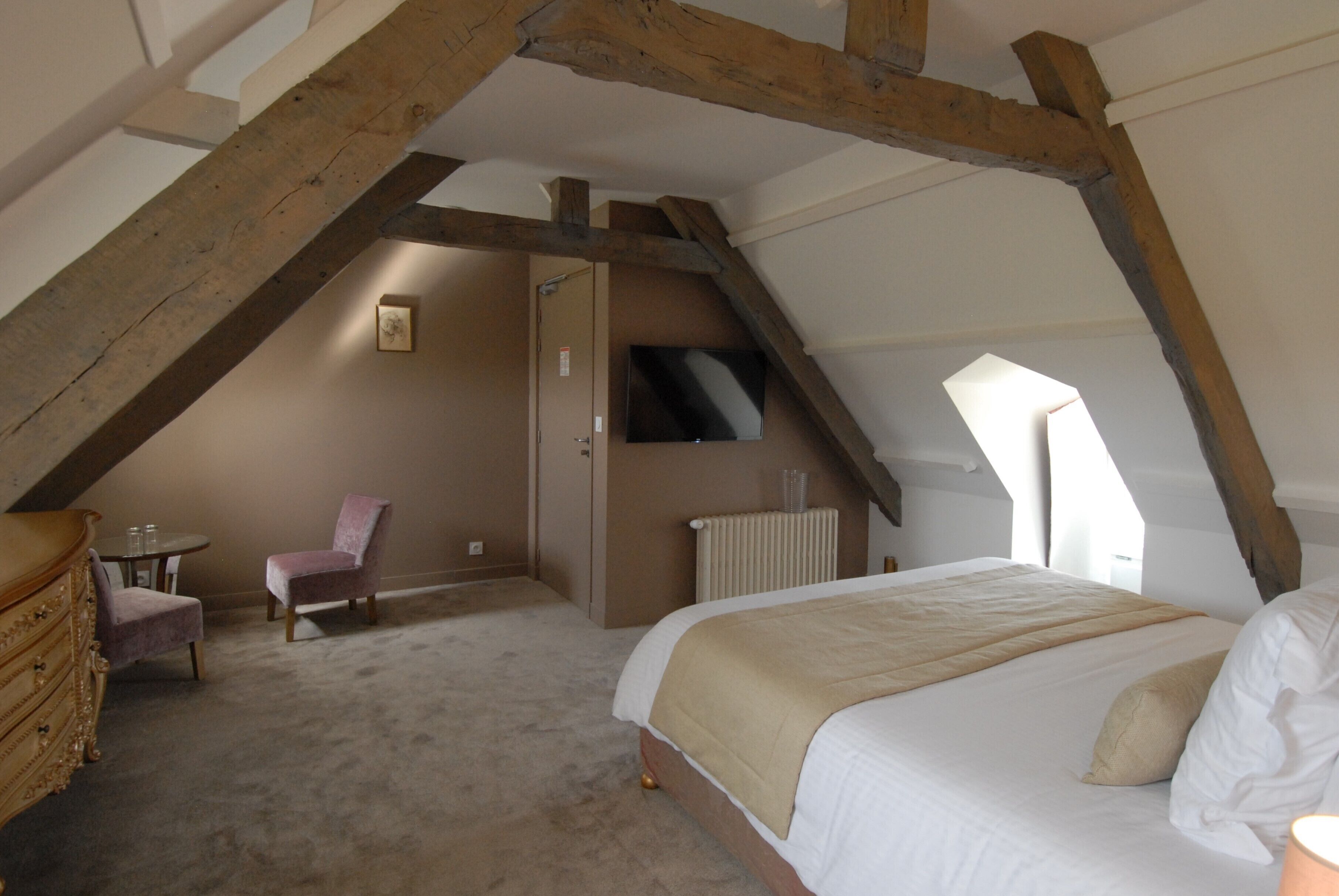Une chambre avec un plafond en pente, un lit, une chaise, une commode et un radiateur.