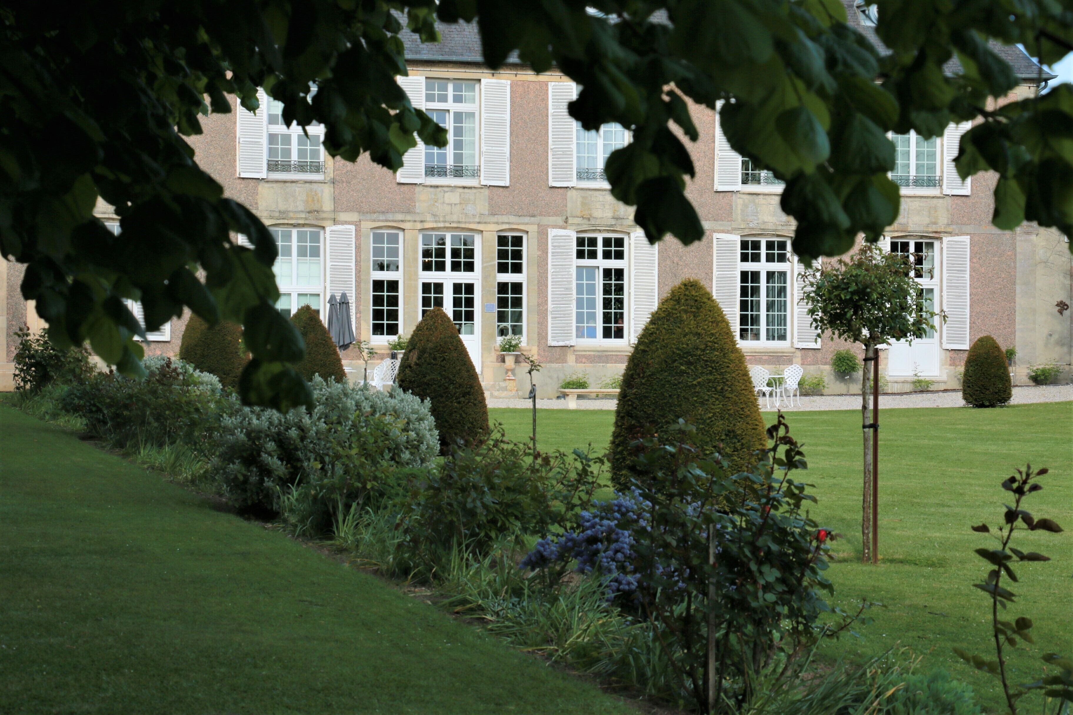 A building with a symmetrical facade, large windows with shutters, and a well-maintained garden with neatly trimmed hedges and shrubs.