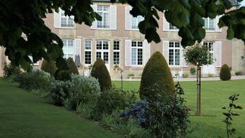 A building with a symmetrical facade, large windows with shutters, and a well-maintained garden with neatly trimmed hedges and shrubs.
