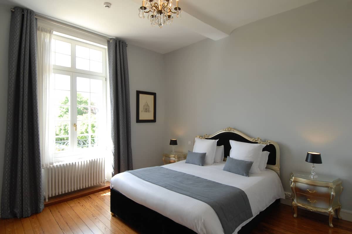 Un dormitorio con una cama grande, un candelabro, una ventana con cortinas y un cuadro en la pared.