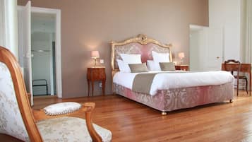A bedroom with a large bed, wooden flooring, a chair, and a nightstand with a lamp.