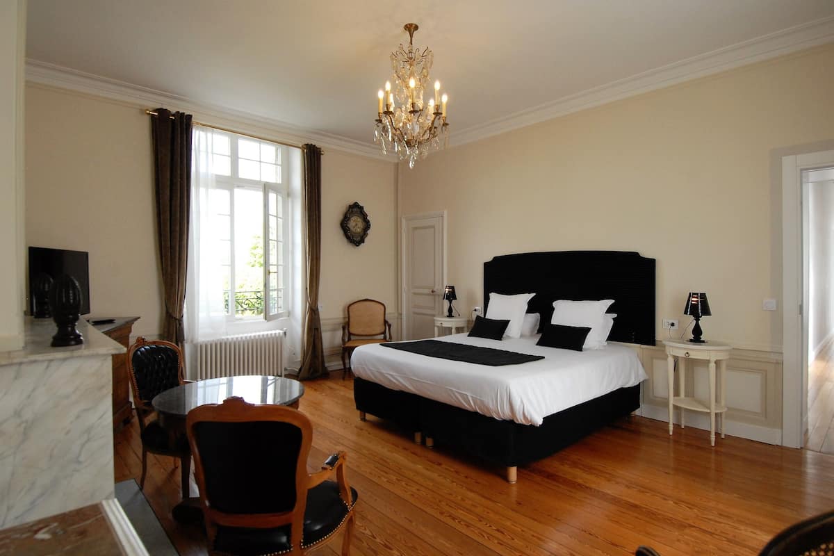 Habitación de hotel con una cama grande, una araña, una mesa de comedor con sillas y vistas al exterior a través de una ventana.