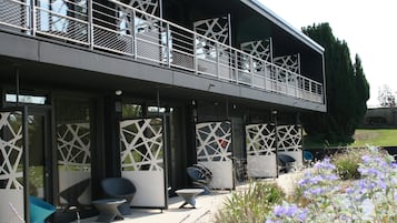 Un edificio moderno con un área de estar al aire libre cubierta, que presenta patrones geométricos en las barandas y un jardín con flores moradas.