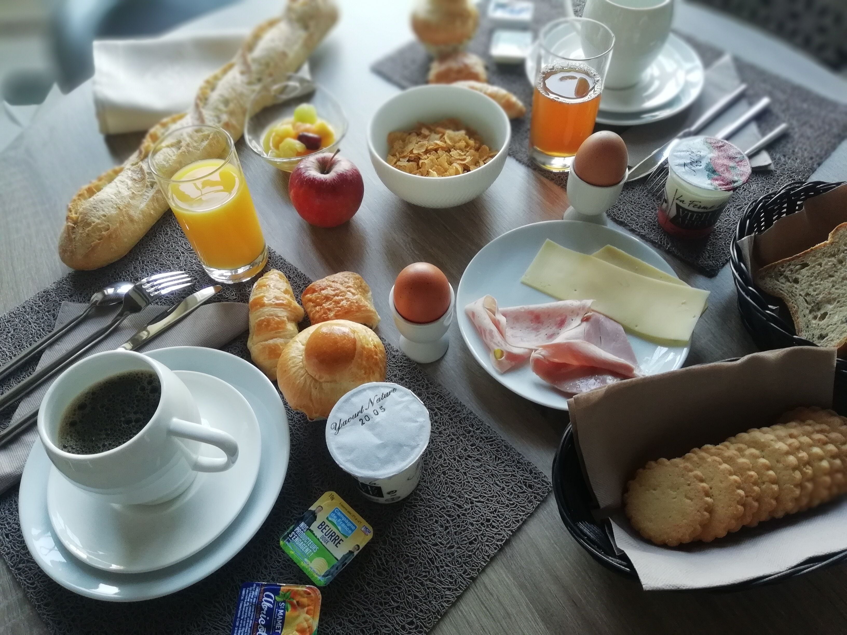 A breakfast table with croissants, a glass of orange juice, a bowl of cereal, a plate of ham and cheese, a cup of coffee, and a small container of yogurt.