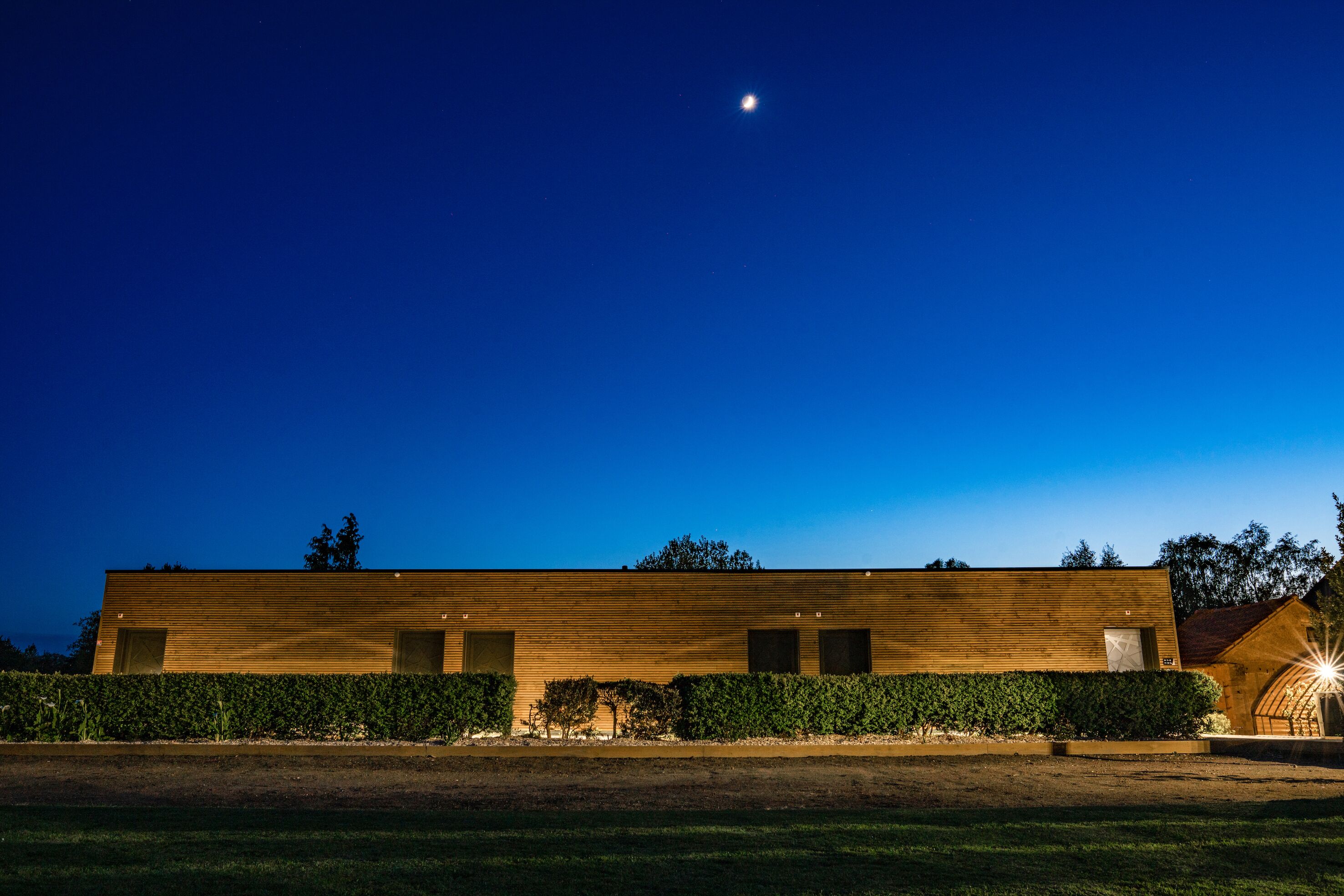 A modern building with a wooden facade, a well-maintained hedge, and a clear night sky with a visible moon.