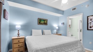 Un dormitorio con una cama, dos mesitas de noche con lámparas, una mesita auxiliar, un cuadro en la pared y una puerta que da a otra habitación.