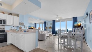 A modern kitchen with white cabinets, a dining area with white furniture, and large windows with blue curtains. - Inn At Summerwind 304 - Hooked on the Beach (Navarre)