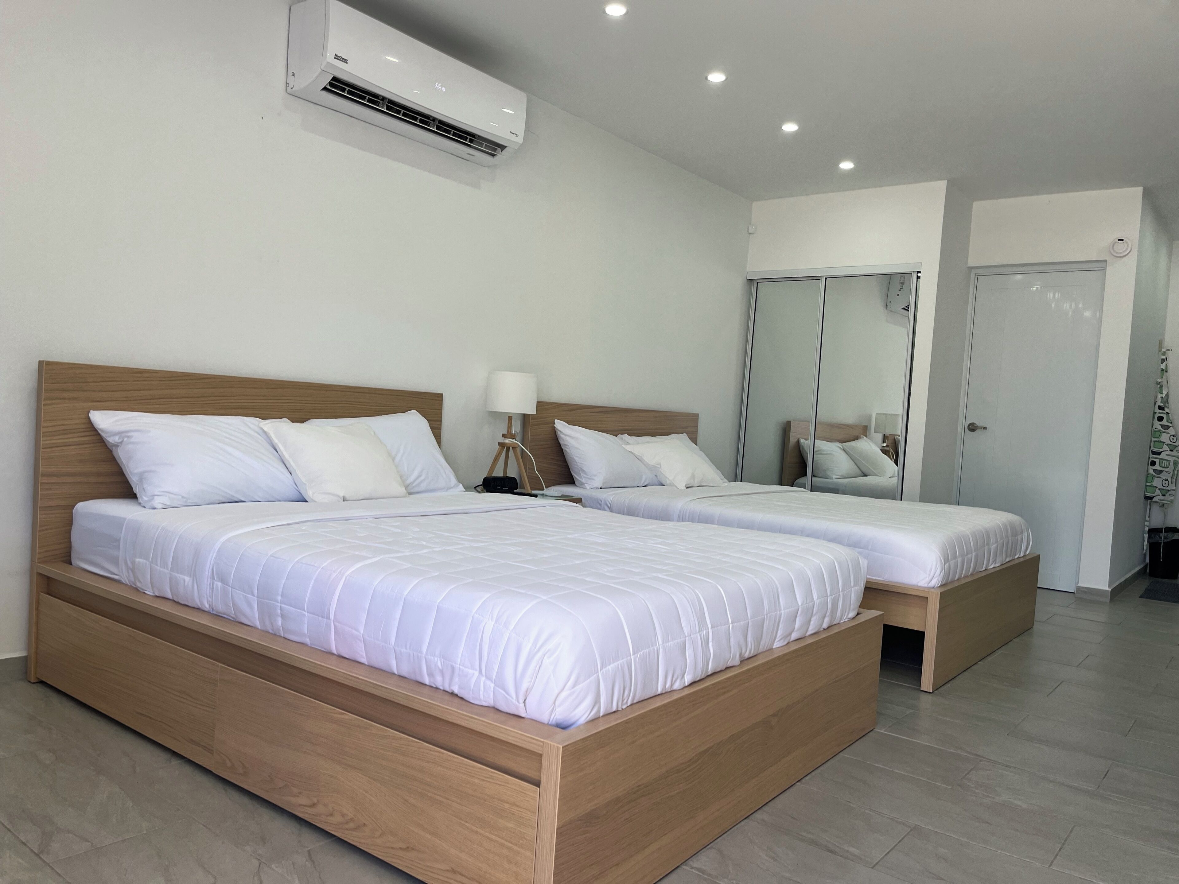 A modern hotel room with two wooden beds, white bedding, a wall-mounted air conditioner, a bedside lamp, and a large mirror.