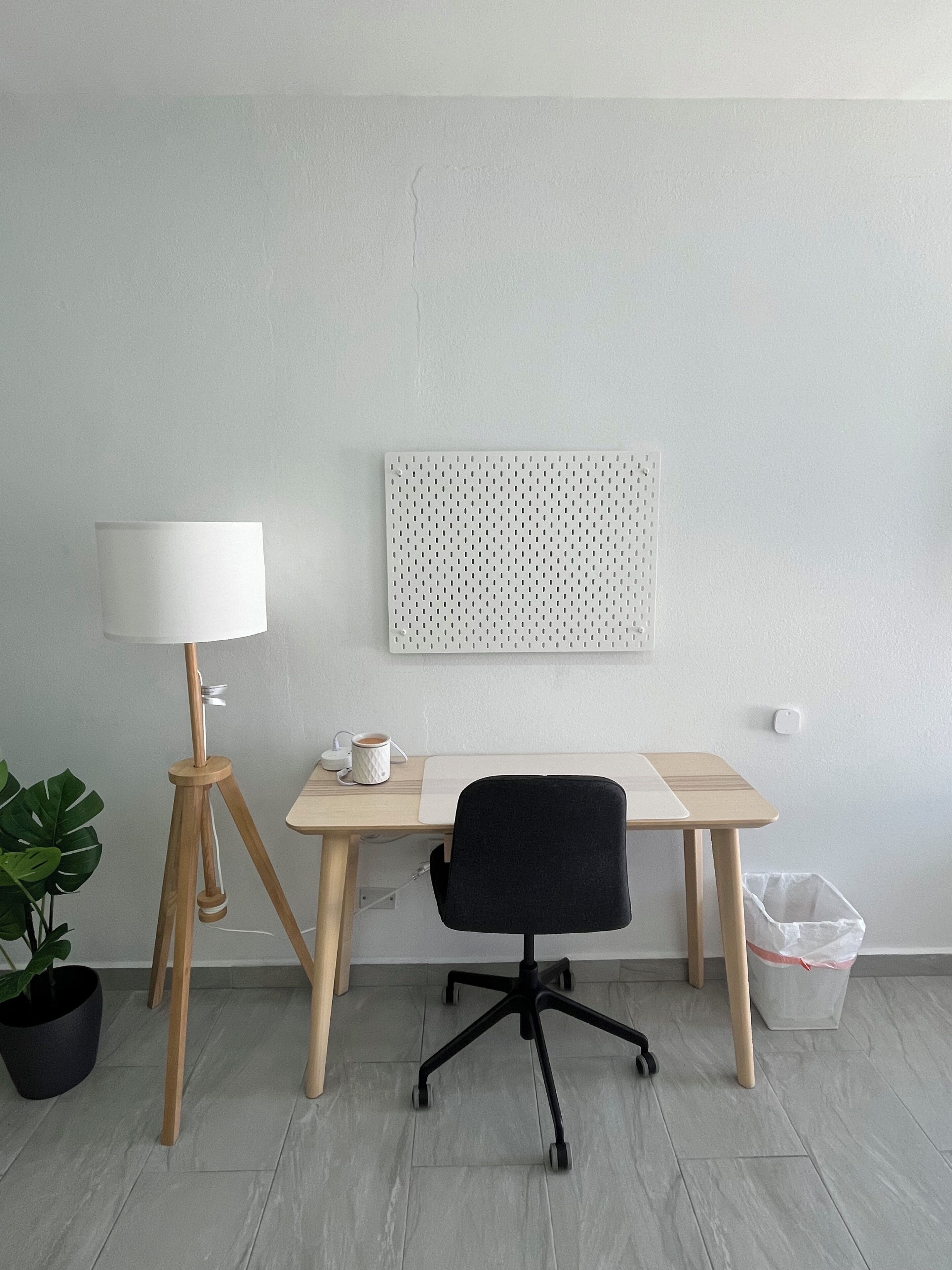 Un espace de travail comprenant un bureau en bois, un fauteuil de bureau noir, un panneau perforé blanc, une lampe sur pied, une tasse à café et une poubelle.