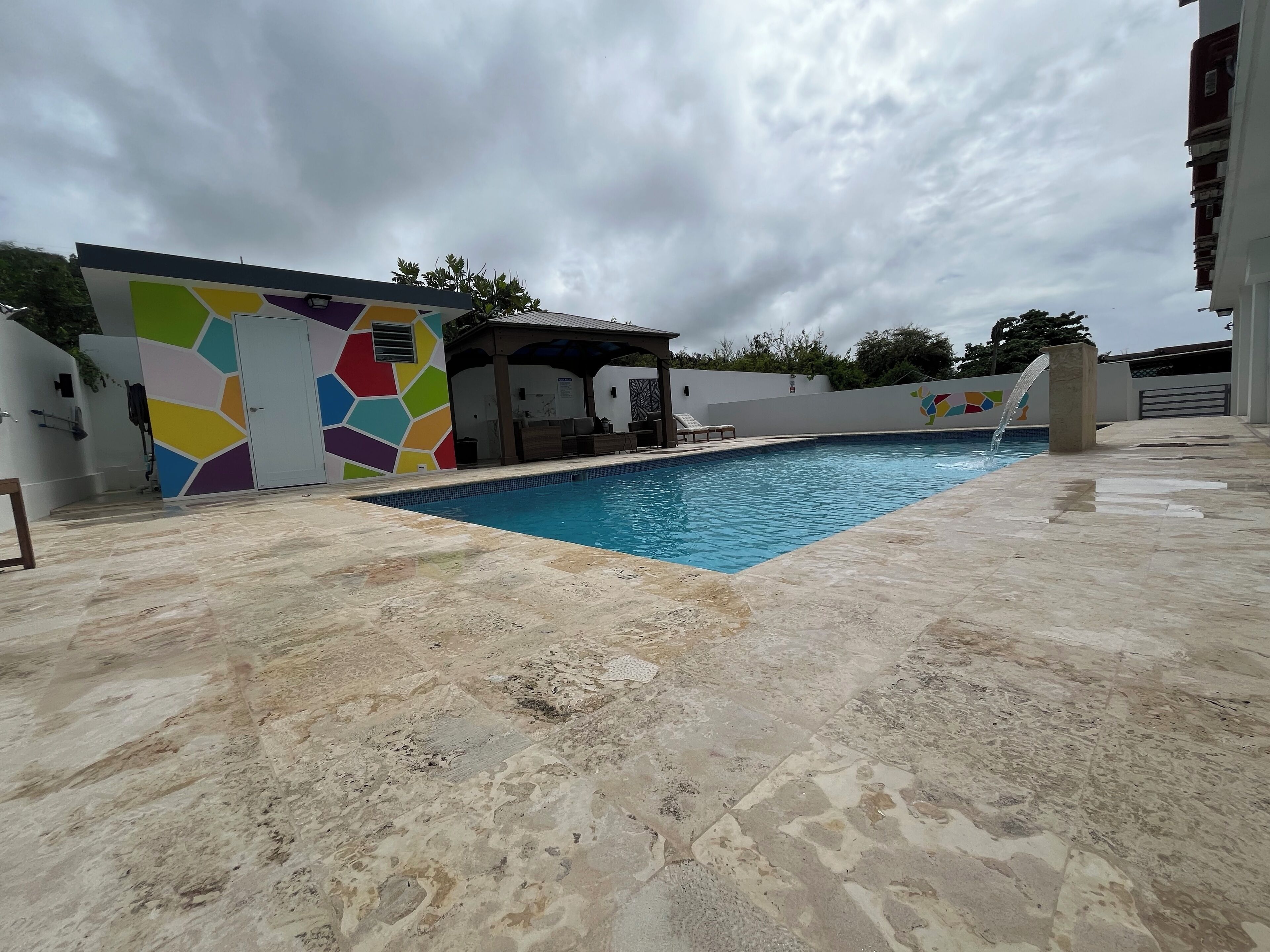 A swimming pool with a tiled deck, a covered seating area, and a colorful wall.