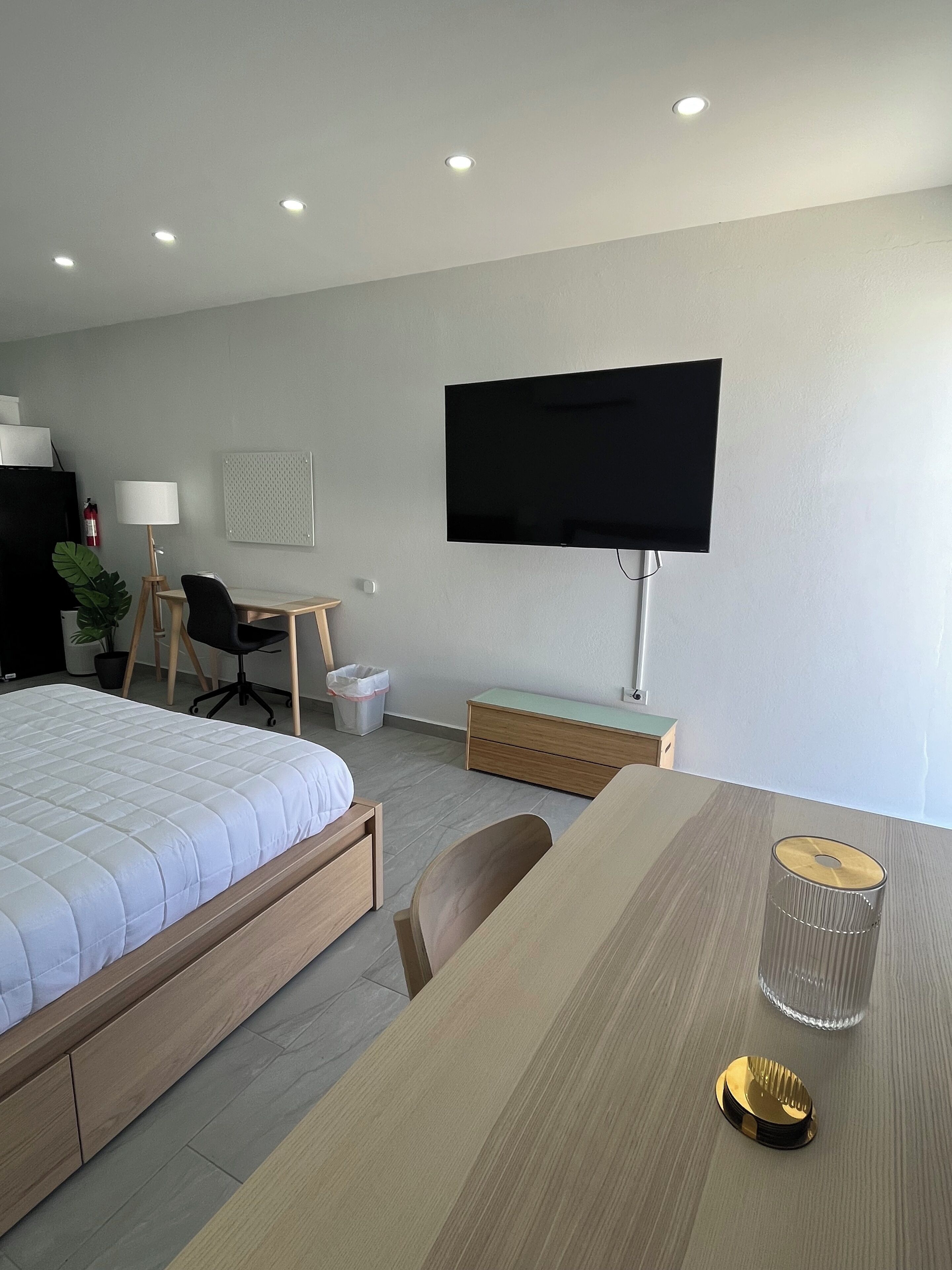 Una habitación de hotel moderna con cama, escritorio, silla, televisor y una mesita con un vaso y una botella.