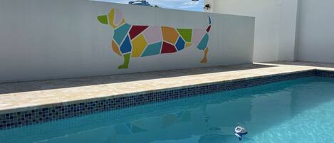 A swimming pool with a tiled edge, a colorful wall mural, and a clear blue sky.