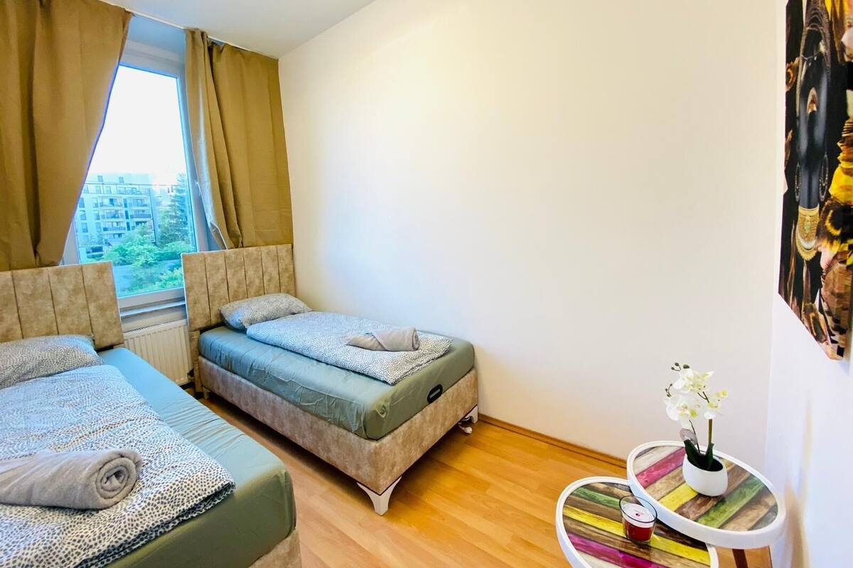A bedroom with two beds, a window with curtains, a small table with a plant, and a colorful wall decoration.