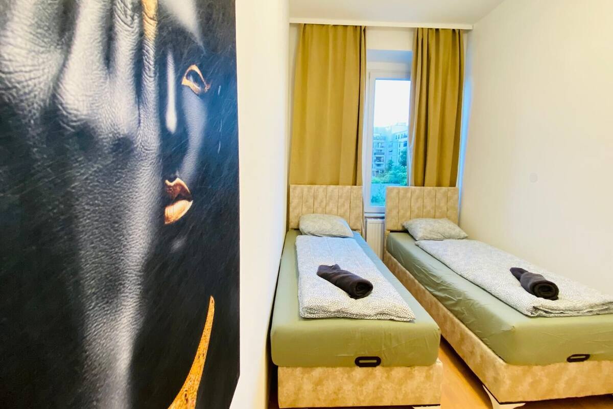 A hotel room with two single beds, a large window with curtains, and a wall with a partial view of a painting.