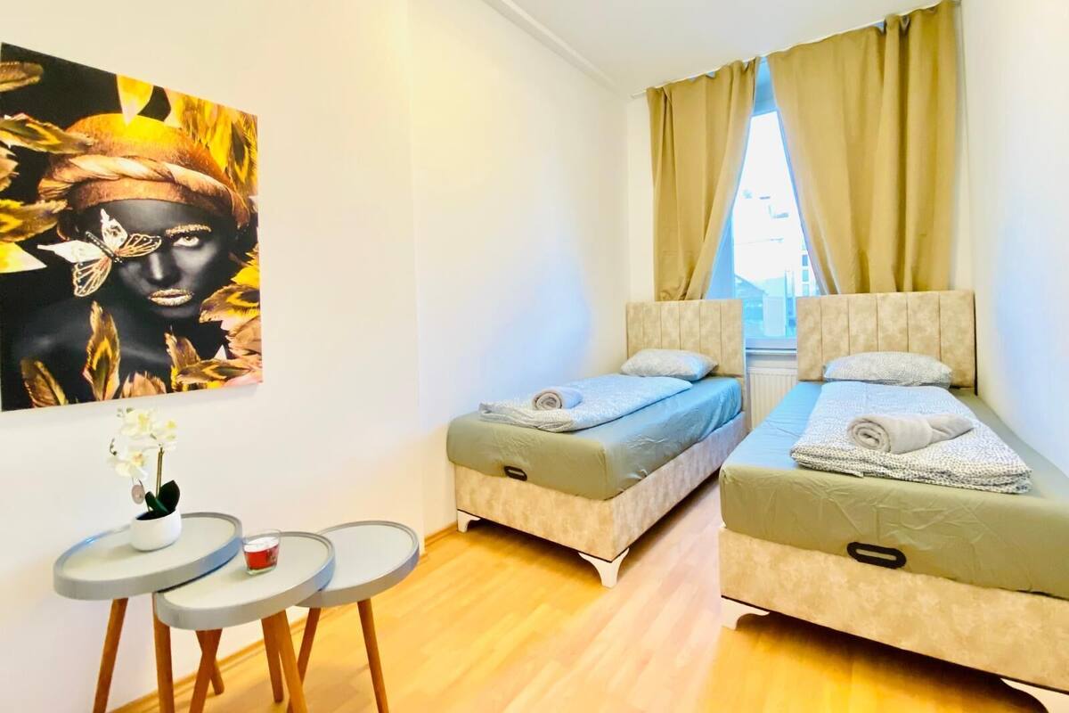 A small, modern room with two single beds, a small table with a vase and candle, and a large window with curtains.