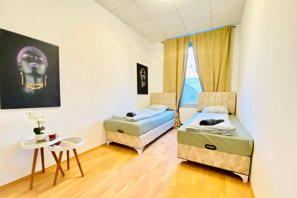 A small, well-lit room with two single beds, a small table with a vase of flowers, and a painting on the wall.
