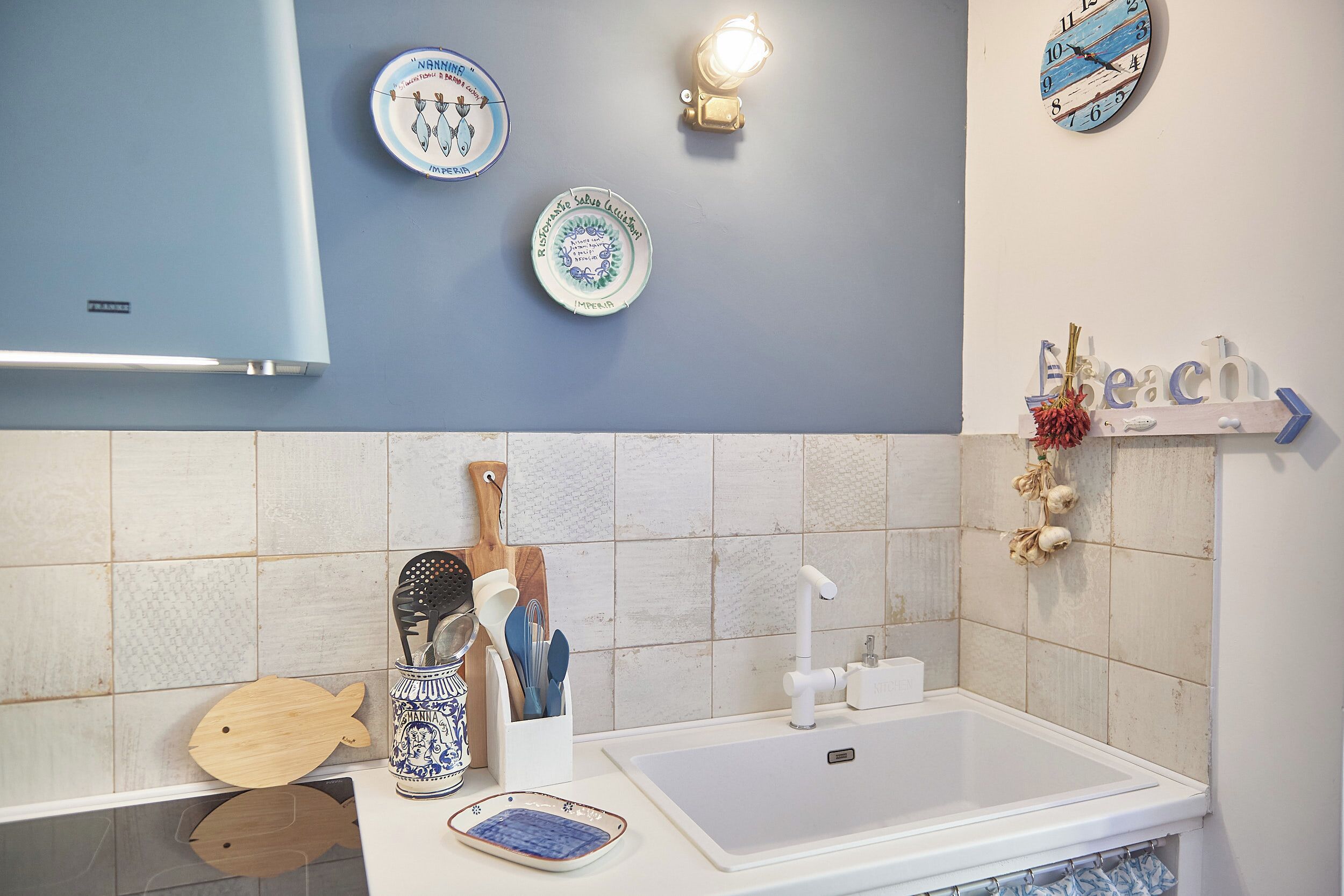Une cuisine avec un plan de travail blanc, un évier et une horloge fixée au mur. On y trouve également des assiettes décoratives et un luminaire mural.