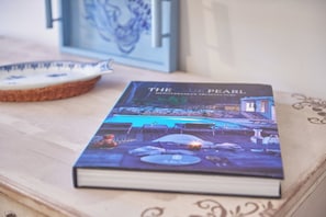 Ein Buch mit dem Titel „The Blue Pearl“ auf einer Holztischplatte, zusammen mit einem dekorativen Teller und einem gerahmten Bild im Hintergrund.