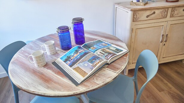 A round wooden table with two white mugs, two blue glass jars, and an open magazine with photos. - La Medusa Blu (Albenga)