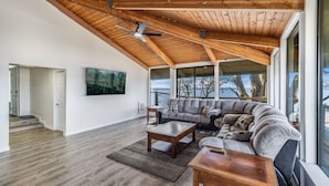 A spacious living room with a high ceiling, wooden beams, a large gray sectional sofa, a wooden coffee table, and a television mounted on the wall. - Dash Point Retreat by the Beach with Amazing Water Views from Almost Every Room. (Tacoma)