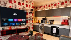 Ett modernt vardagsrum med en platt-TV som visar TV-kanaler, en soffa, ett soffbord och en köksdel med vitvaror.