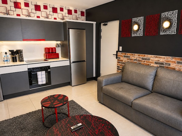 Cocina moderna con refrigerador, estufa y electrodomésticos rojos. Hay un sofá gris y una pequeña mesa redonda con diseño rojo y negro.