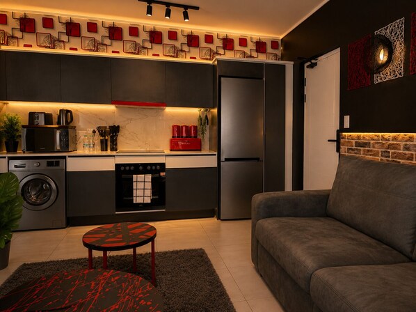 A modern kitchen with a built-in oven, microwave, and refrigerator. There is a grey sofa and a small round table with a red and black design.