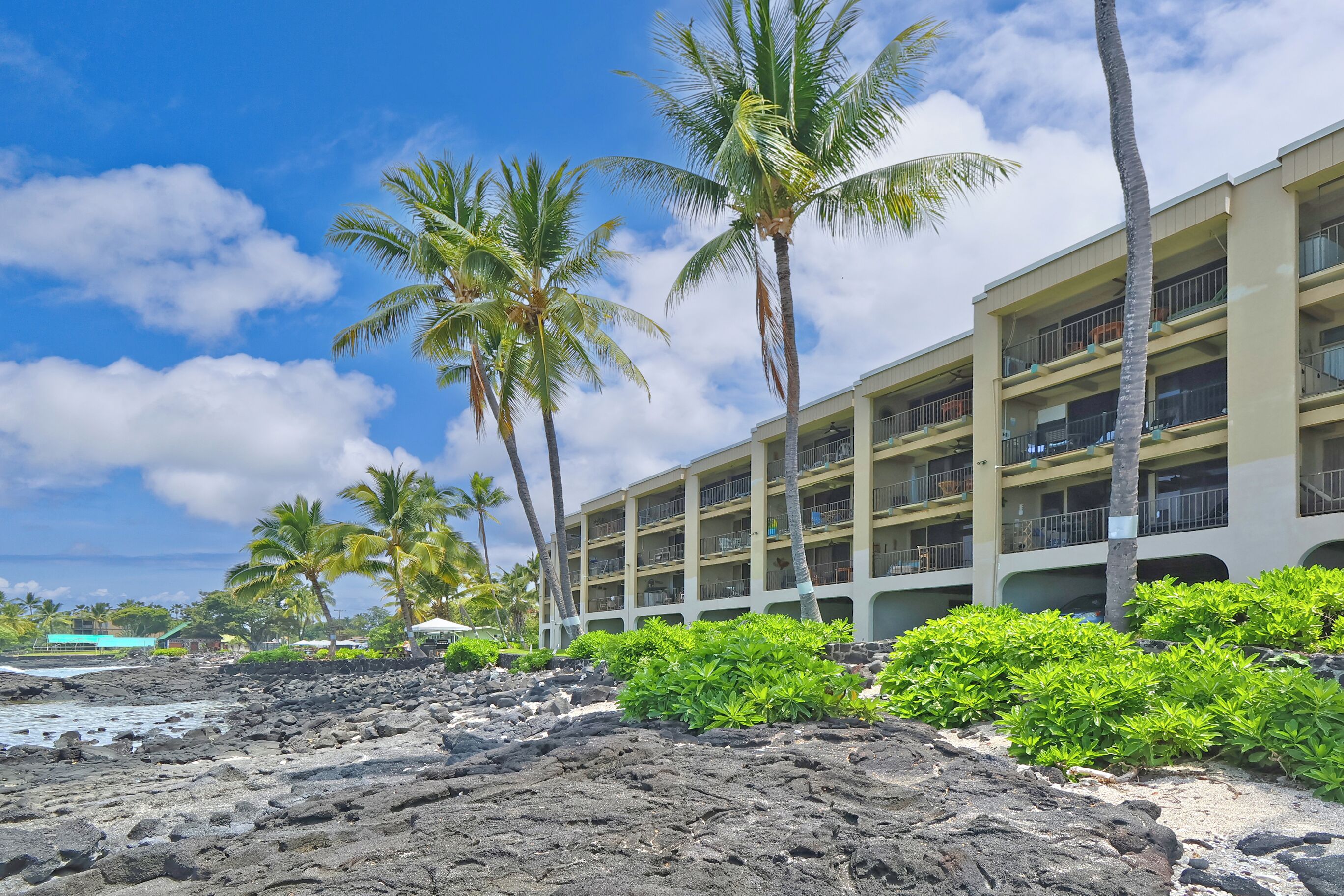 A hotel with multiple balconies, a rocky beach, and palm trees.