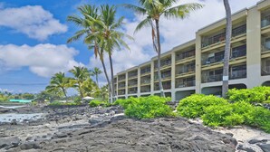 A hotel with multiple balconies, a rocky beach, and palm trees.