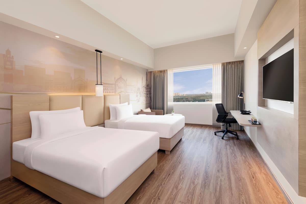 Habitación de hotel con dos camas, un escritorio, una silla, un televisor y una ventana con vistas a la ciudad.