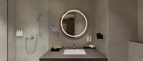 A modern bathroom with a granite countertop, a round mirror, a handheld showerhead, and a towel rack.