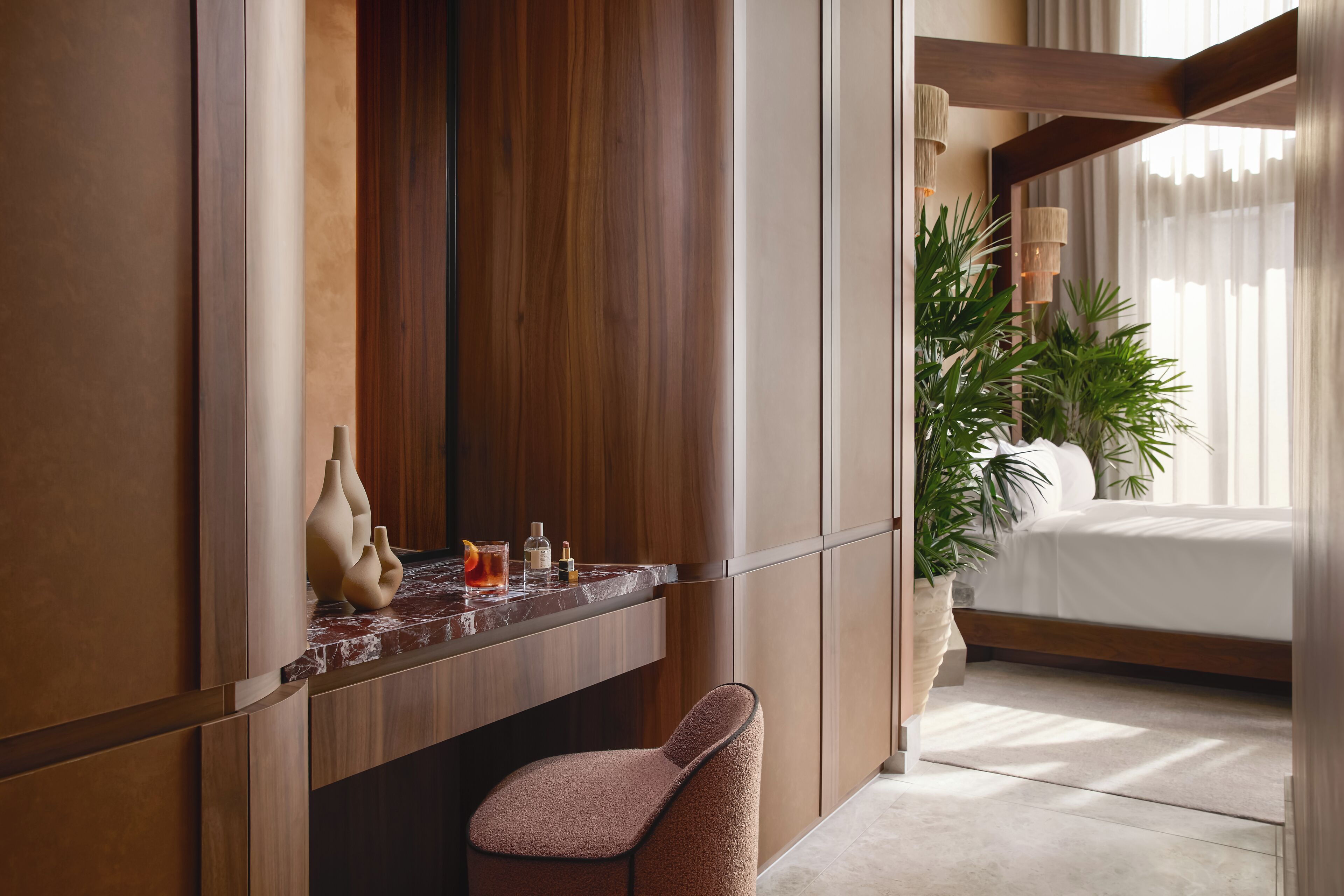 A modern hotel room with a wooden vanity, a marble countertop, a plush chair, a large potted plant, and a neatly made bed.