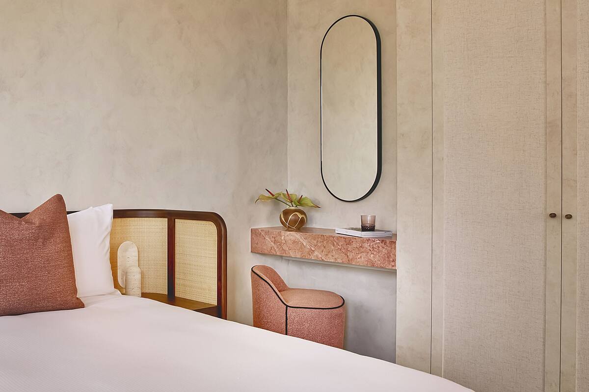 A bedroom with a bed, a nightstand, a mirror, a small table with a vase and a book, and a door.