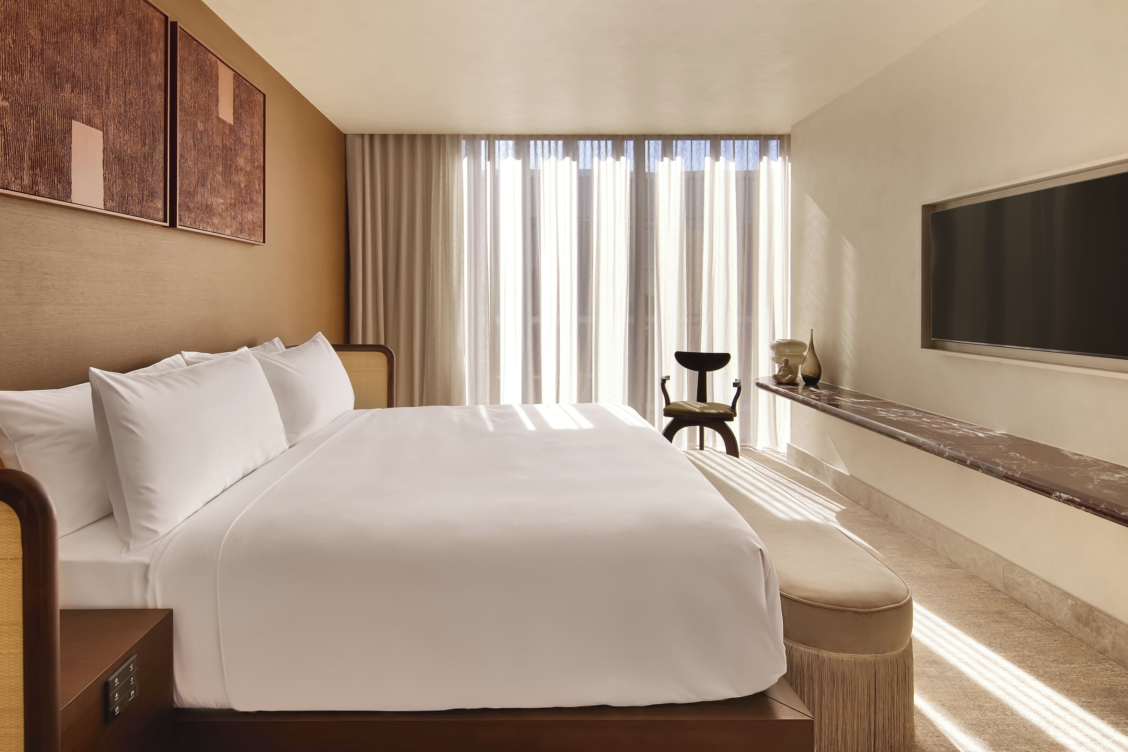 Une chambre d’hôtel avec un grand lit, un téléviseur fixé au mur, un bureau avec une lampe et une vue sur l’extérieur à travers des rideaux transparents.