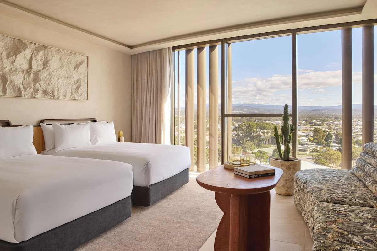 A hotel room with two beds, a sofa, a round wooden table, and a large window with a city view.