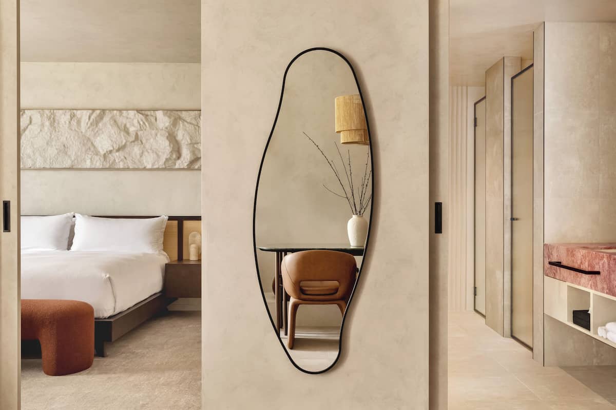 A modern hotel room with a large mirror, a bed, a chair, a nightstand, and a bathroom with a marble countertop.