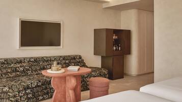 A hotel room with a sofa, a round wooden table with a book and a tray, a cabinet with bottles, and a bed.