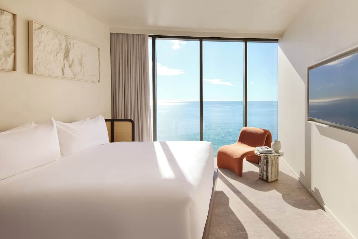 A hotel room with a large bed, a view of the ocean, a small orange chair, and a round side table with a vase and books.