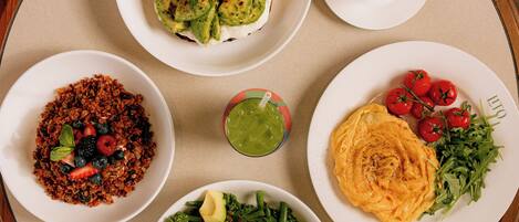 A round table with various dishes including a salad, a plate with cherry tomatoes, a bowl of granola, a plate with avocado and greens, and a cup of coffee.