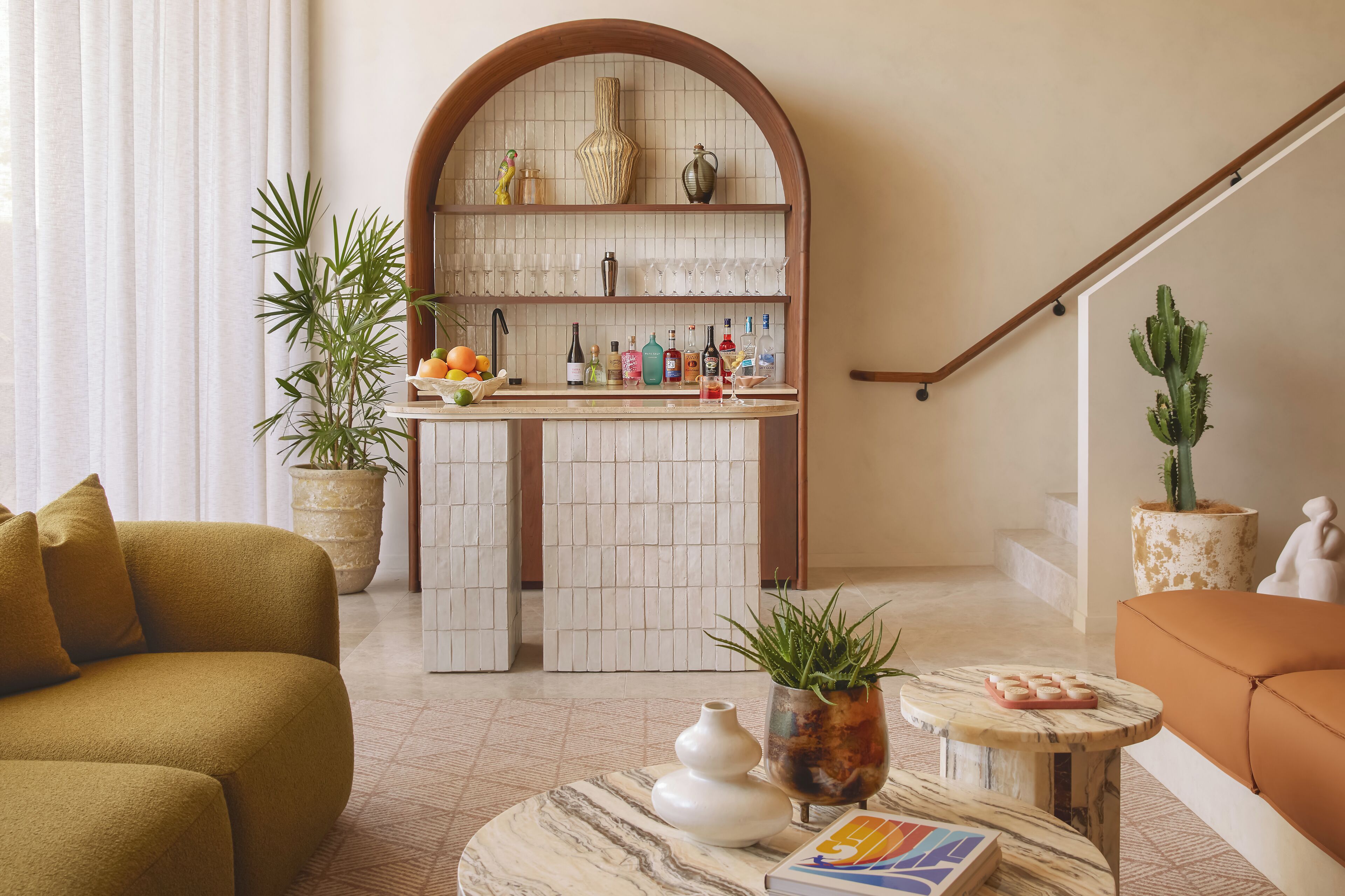 A modern living room with a kitchen area, featuring a curved wooden arch, a marble coffee table, and a variety of plants.