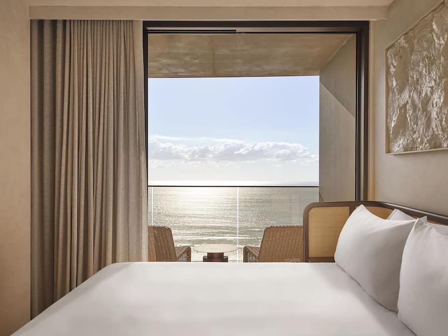 A hotel room with a bed, a view of the sea, and a large window with curtains.