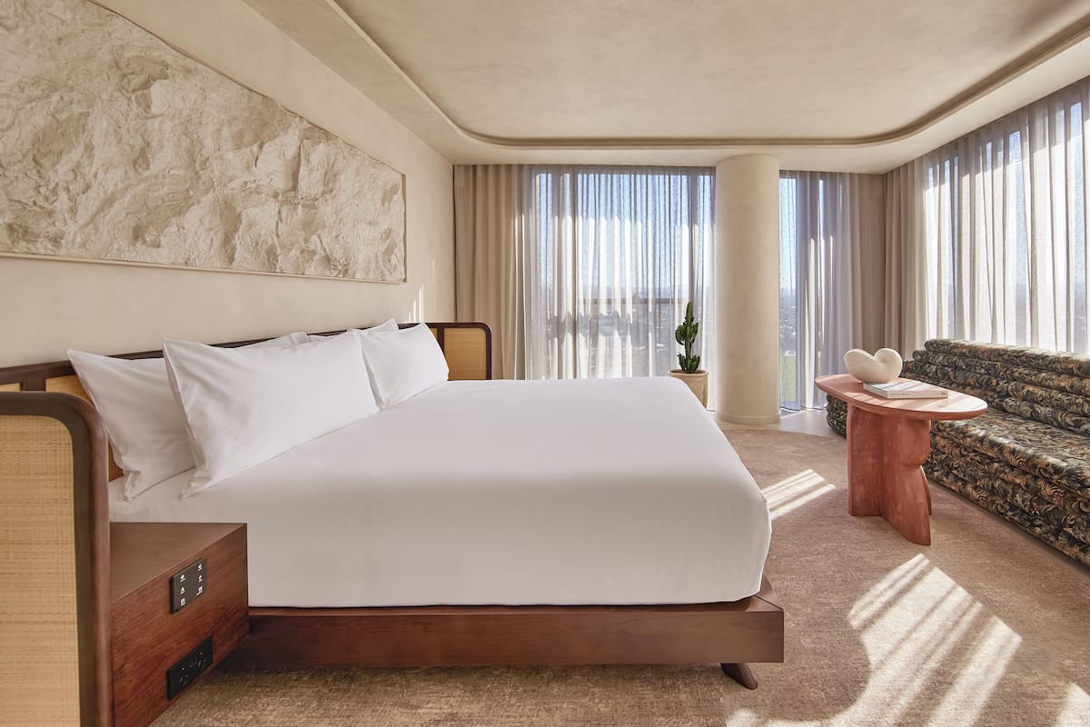 A modern hotel room with a large bed, a sofa, a small table, and a large window with curtains.