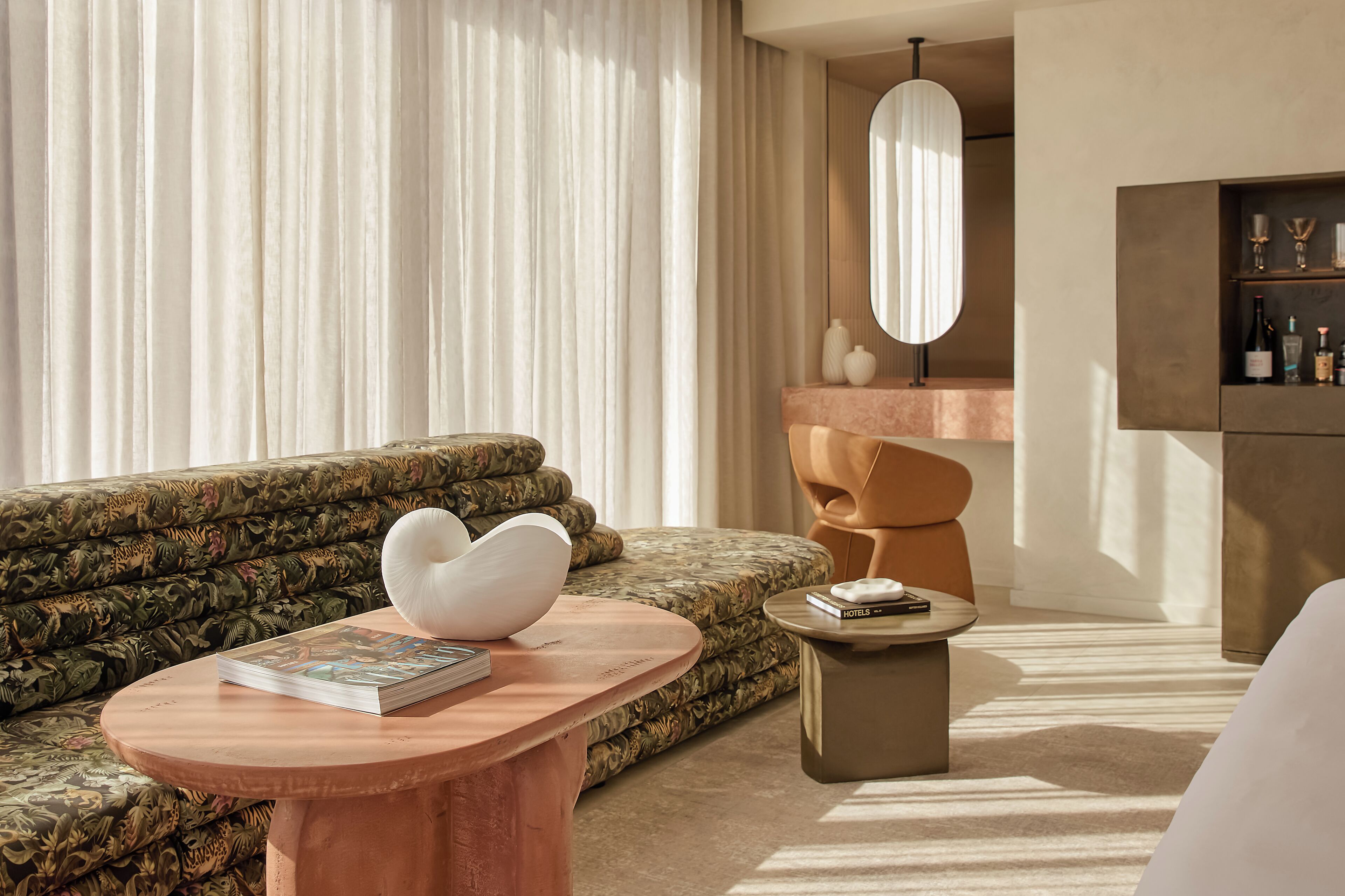 A modern hotel room with a patterned sofa, a round wooden coffee table, a small side table, a large mirror, and a minibar.